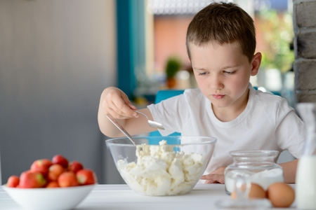 7 year old boy adding sugar to cottage cheese in a bowl. Prepares mini cheesecakes with strawberriesの写真素材