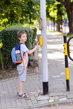 Little 7 years schoolboy pressing a button on traffic lights and waiting for green light. Dressed in white t shirt and shorts. Blue backpack with watercolors paintsの写真素材