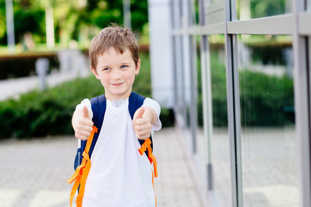 Happy little 7 years schoolboy showing thumbs up gesture. Dressed in white t shirt. Blue backpackの写真素材