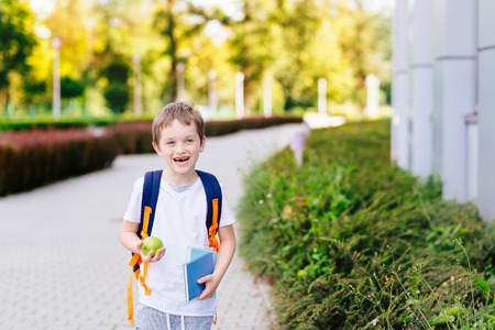 Happy little 7 years old boy at his first day at school . Dressed in white t shirt. Boy holding apple and blue book. Blue backpackの写真素材