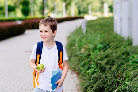 Happy little 7 years old boy at his first day at school . Dressed in white t shirt. Boy holding apple and blue book. Blue backpackの写真素材