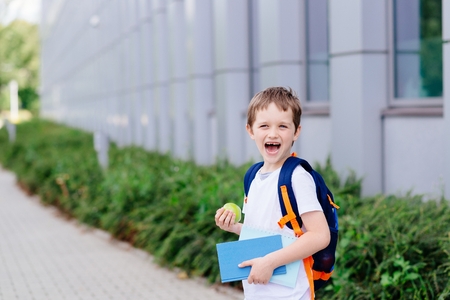 Happy little 7 years old boy at his first day at school . Dressed in white t shirt. Boy holding apple and blue book. Blue backpackの写真素材