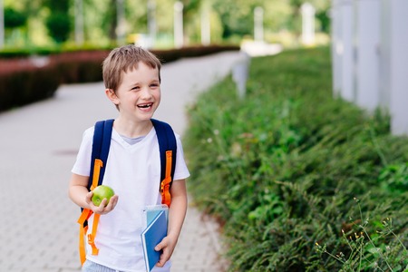 Happy little 7 years old boy at his first day at school . Dressed in white t shirt. Boy holding apple and blue book. Blue backpackの写真素材
