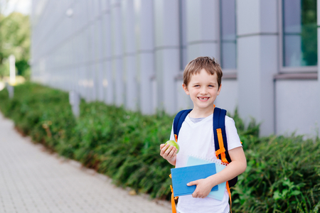 Happy little 7 years old boy at his first day at school . Dressed in white t shirt. Boy holding apple and blue book. Blue backpackの写真素材