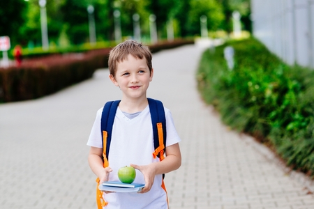 Little 7 years old boy with books and apple at road to school. Dressed in white t shirt and shortsの写真素材