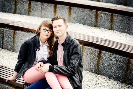 Young couple in love hugging on the bench. Engagement session. Dressed in black leather jackets. Girl wears glassesの写真素材