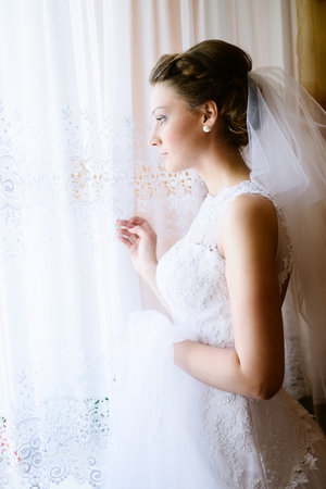 Young bride in white dress looking out the window. Wedding preparations. Bridal morning of a cute lady. Wedding day.の写真素材