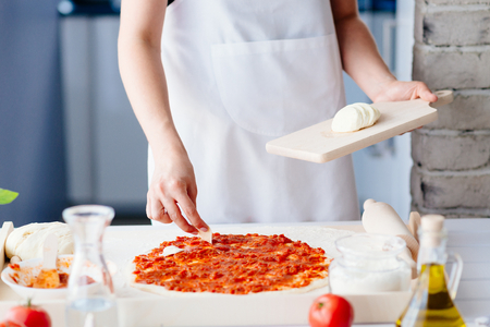 Woman puts slices of mozzarella on the pizza. Woman doing homemade pizza. Dressed in white pinnyの写真素材