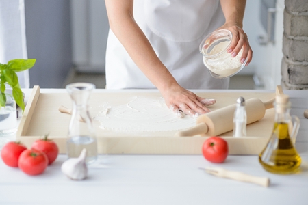 Woman sprinkling wooden pastry board with flour. Woman doing homemade pizza. Dressed in white pinnyの写真素材