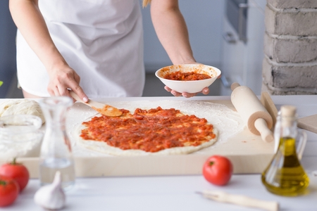 Woman adding tomato sauce on pizza. Woman doing homemade pizza. Dressed in white pinnyの写真素材