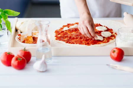 Woman puts slices of mozzarella on the pizza. Woman doing homemade pizza. Dressed in white pinnyの写真素材