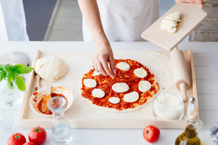 Woman puts slices of mozzarella on the pizza. Woman doing homemade pizza. Dressed in white pinnyの写真素材