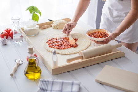 Woman adding tomato sauce on pizza. Woman doing homemade pizza. Dressed in white pinnyの写真素材