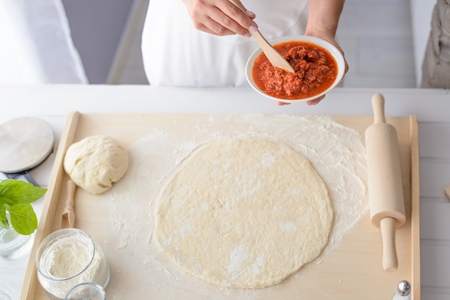 Woman adding tomato sauce on pizza. Woman doing homemade pizza. Dressed in white pinnyの写真素材