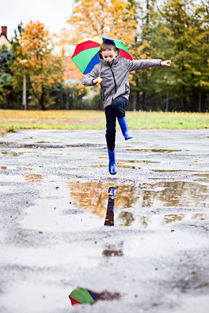 Happy little boy in rubber blue rainboots jumping to dirty puddle. Rainy autumn dayの写真素材