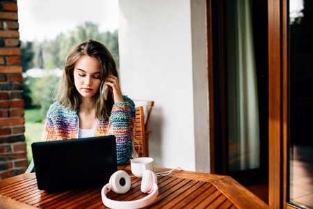 Young woman in woolen sweater uses a smartphone and laptop computer on home terrace.の写真素材