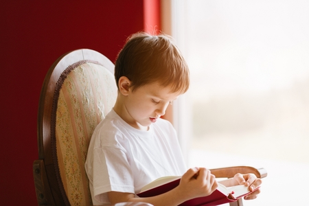 Little 7 years old boy sitting on old armchair and reading a book.の写真素材