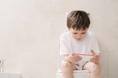Little 7 years old boy playing with smartphone while sitting on toilet.の写真素材