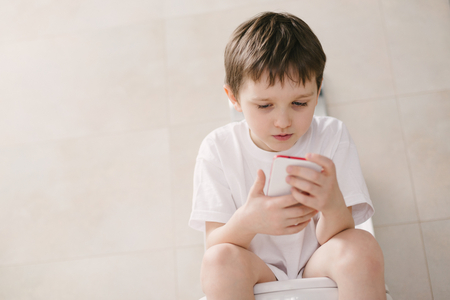 Little 7 years old boy playing with smartphone while sitting on toilet.の写真素材