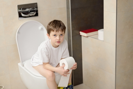Little 7 years old boy sitting on toilet. Holding white toilet paperの写真素材