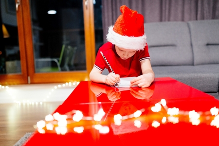 Pensive little boy writing a letter to Santa Claus. Wears red Santa Claus hat and red polo shirtの写真素材