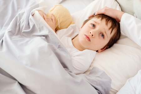 Sick little child with temperature in bed. Hand of doctor on his foreheadの写真素材