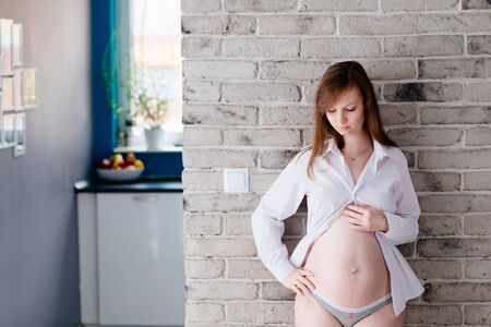 Beautiful young pregnant woman posing by the grey brick wall in white shirtの写真素材