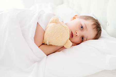 Little boy child sleepy in bed. Hugging his favourite teddy bear. Covered with white duvetの写真素材