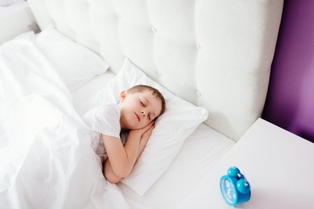 Little boy child sleeping in bed. Hugging his favourite teddy bear. Covered with white duvet. Blue alarm clock on bedside cabinet. Its 7amの写真素材