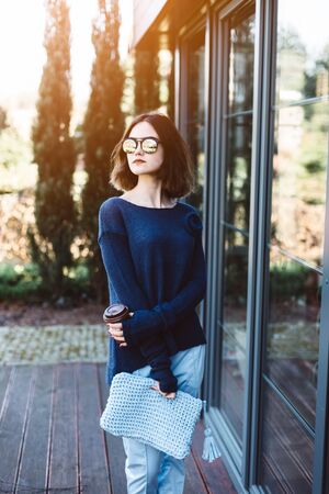 Young girl holding paper mug with hot coffee on terrace. Wearing blue woolen sweater and sunglassesの写真素材