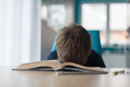 Tired 8 years old boy doing his homework at the table. Child reading a book at the desk.