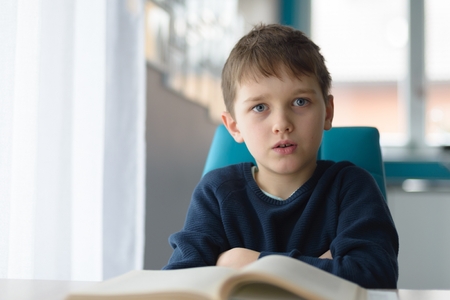 Tired 8 years old boy doing his homework at the table. Child reading a book at the desk.の写真素材