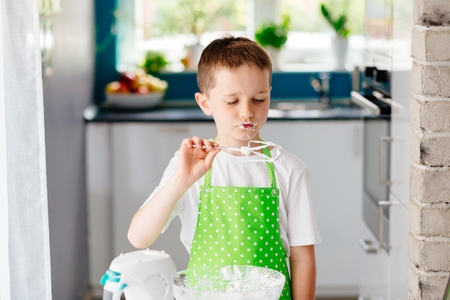 Happy child trying sweet dough from the mixer beater after mixing for cake. Child helping in kitchenの写真素材