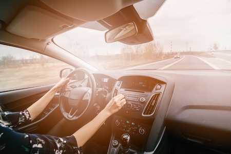 Young woman driving a car and adjusting car audio. Music in a car. Sunny day. Modern car interiorの写真素材