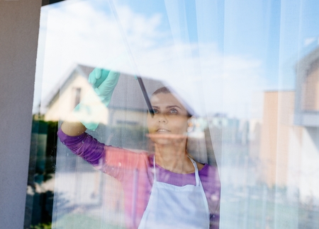 Young beautiful woman in white apron cleaing windows. Cleaning service. Maid cleaning at homeの写真素材