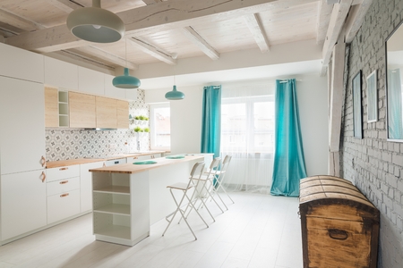 Open bright kitchen with white furniture. Island kitchen. Turquoise lamps and curtains. Modern, bright attic apartment.の写真素材