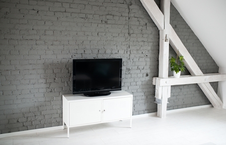 Modern flat lcd television set on white cabinet. Grey brick wall and wooden ceiling beams. Modern, bright attic apartment.の写真素材