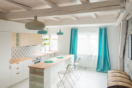 Open bright kitchen with white furniture. Island kitchen. Turquoise lamps and curtains. Modern, bright attic apartment.の写真素材