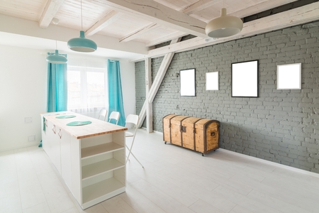 Gray brick wall with empty frames. Open bright kitchen with white furniture. Island kitchen. Turquoise lamps and curtains. Modern, bright attic apartment.の写真素材