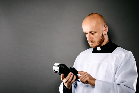 Priest in white surplice and black shirt with white collar holding credit card terminal. Church and money conceptの写真素材