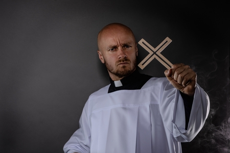 Catholic priest in white surplice and black shirt with cleric collar holding wooden cross and prayingの写真素材