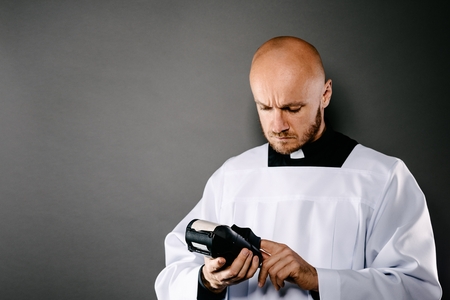 Priest in white surplice and black shirt with white collar holding credit card terminal. Church and money conceptの写真素材