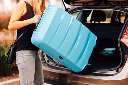 Young woman loading two blue plastic suitcases to car trunk. Preparation for tripの写真素材