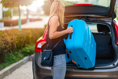 Young woman loading two blue plastic suitcases to car trunk. Preparation for tripの写真素材