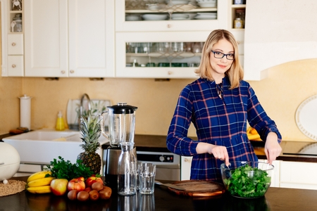 Woman mixing green kale in a glass bowl in kitchen. Woman in kitchen. Female preparing delicious healthy fruit and vegetable smoothieの写真素材