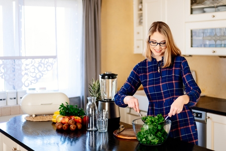 Woman mixing green kale in a glass bowl in kitchen. Woman in kitchen. Female preparing delicious healthy fruit and vegetable smoothieの写真素材