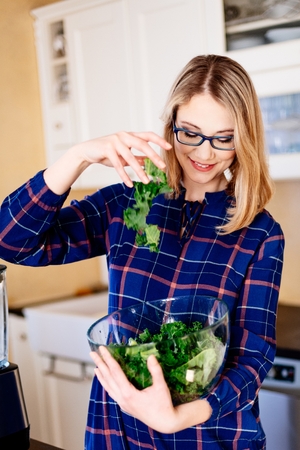 Young pregnant woman mixing green kale in a glass bowl in kitchen. Woman in kitchen. Female preparing delicious healthy fruit and vegetable smoothieの写真素材