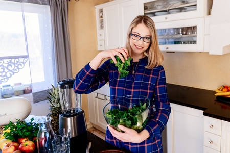 Young pregnant woman mixing green kale in a glass bowl in kitchen. Woman in kitchen. Female preparing delicious healthy fruit and vegetable smoothieの写真素材