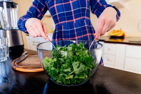 Woman mixing green kale in a glass bowl in kitchen. Woman in kitchen. Female preparing delicious healthy fruit and vegetable smoothieの写真素材
