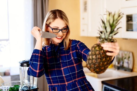 Young woman opening pineapple with knife in kitchen. Woman in kitchen. Female preparing delicious healthy fruit and vegetable smoothieの写真素材